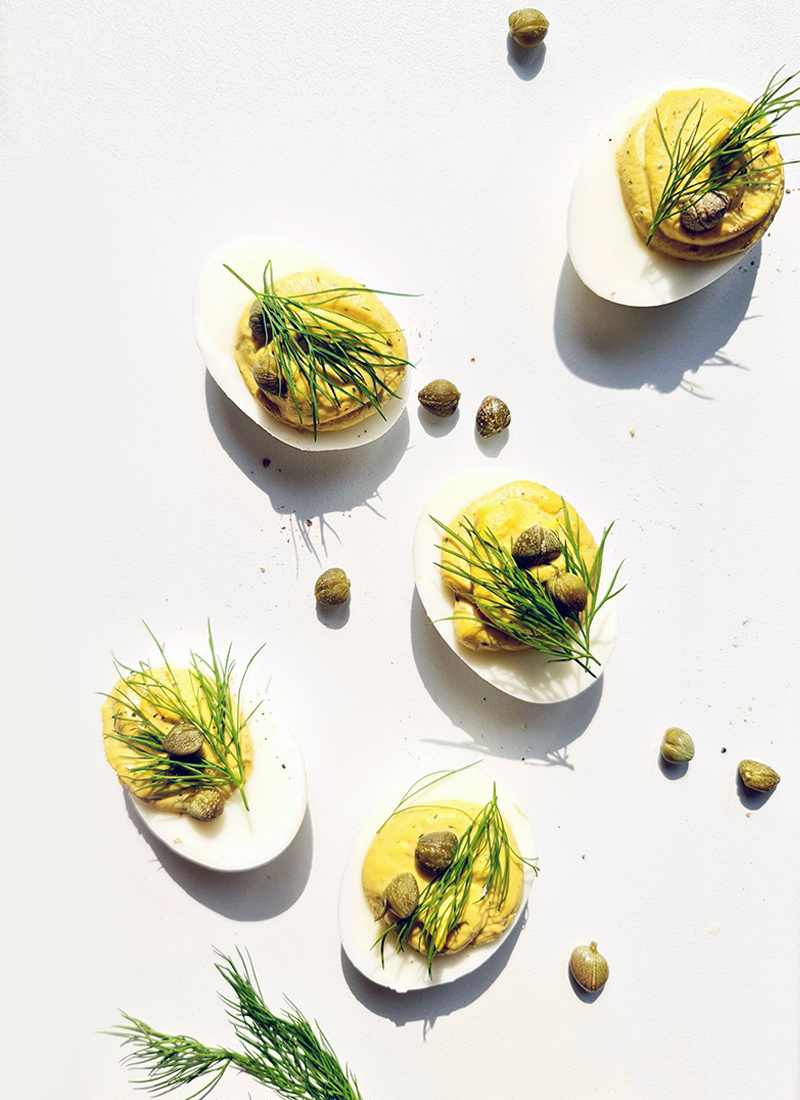 Gourmet deviled eggs with tuna and capers, garnished with dill, cast in soft light on a white surface.