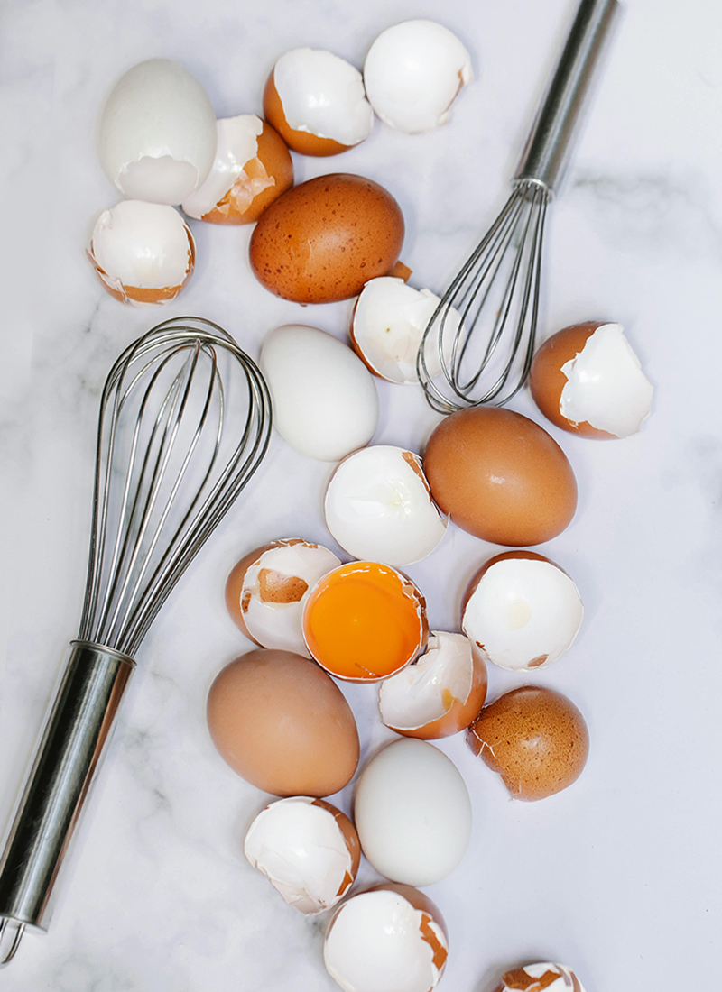 Eggs, both brown and white, scattered on a marble surface with cracked shells and egg yolk visible. Two stainless steel whisks lie amidst the eggs, perfect for quick cooking hacks to save dinner.