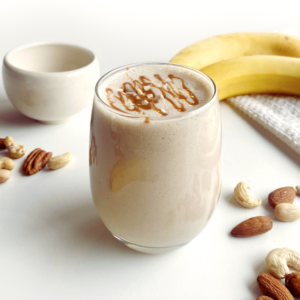 A Peanut Butter Banana Smoothie in a glass decorated with chopped almonds, surrounded by bananas, nuts, and a small bowl on a white surface.