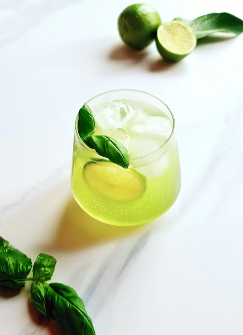 A glass of lime basil mocktail, perfect as a drink for any occasion, with ice cubes, garnished with fresh basil leaves on a white marble surface, accompanied by limes.