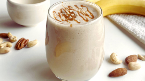 A Peanut Butter Banana Smoothie in a glass decorated with chopped almonds, surrounded by bananas, nuts, and a small bowl on a white surface.