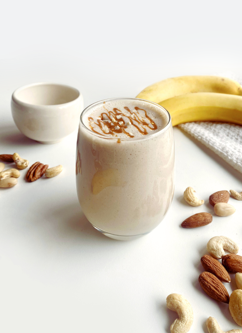 A Peanut Butter Banana Smoothie in a glass decorated with chopped almonds, surrounded by bananas, nuts, and a small bowl on a white surface.