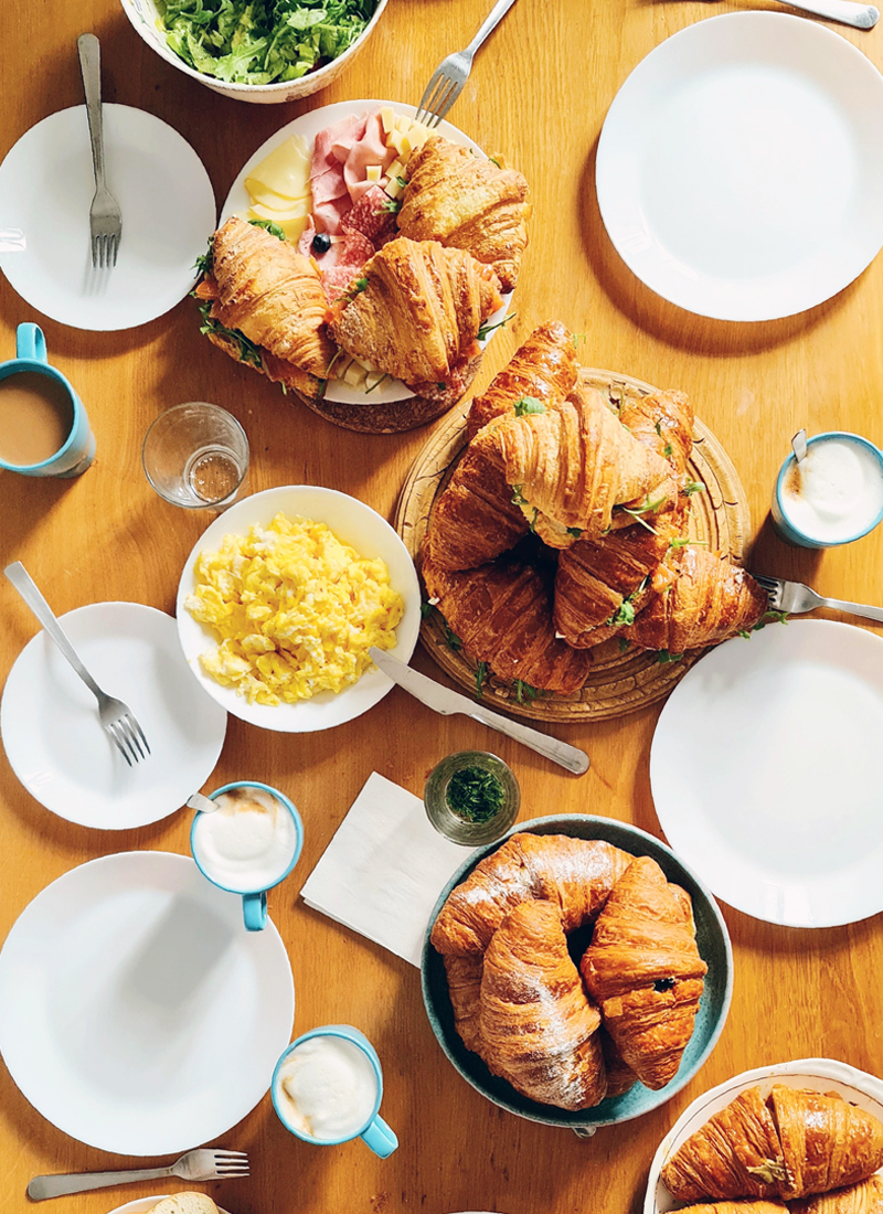 A breakfast spread on a wooden table featuring croissants, some filled with salmon, arugula and cheese, scrambled eggs, cups of yogurt, and empty plates awaiting guests.