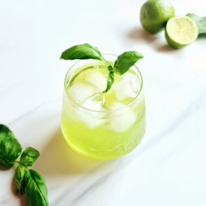 A glass of lime basil mocktail, perfect as a drink for any occasion, with ice cubes, garnished with fresh basil leaves on a white marble surface, accompanied by limes.