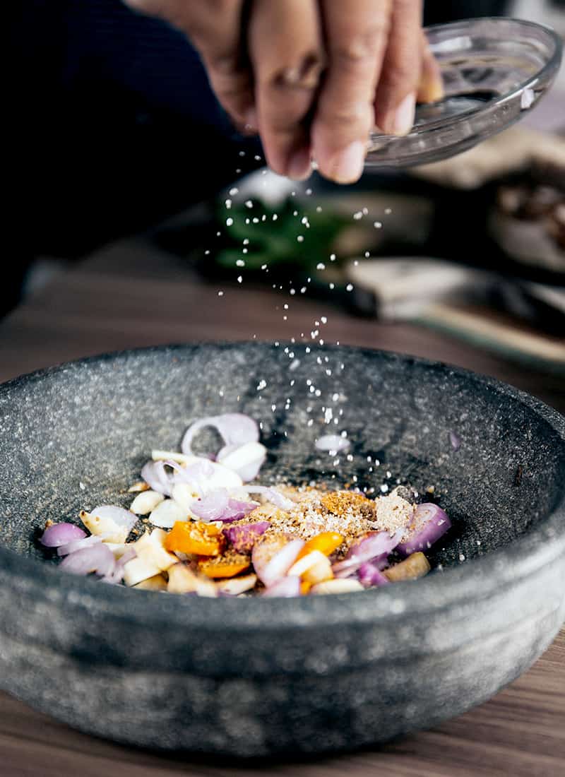 A hand sprinkling salt over a bowl containing a colorful mix of spices, herbs, and chopped ingredients, suggesting the preparation of a fresh, aromatic spice blend or dish.