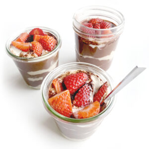 Three glasses of layered overnight oatmeal with fresh strawberries, creamy filling, and a high-protein chocolate-flavored layer, served with a spoon, on a white background.