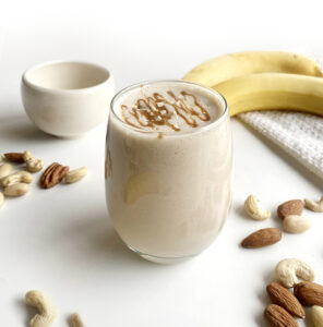 A Peanut Butter Banana Smoothie in a glass decorated with chopped almonds, surrounded by bananas, nuts, and a small bowl on a white surface.