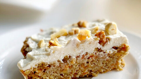 A slice of healthy low calorie carrot cake topped with chopped nuts, served on a white plate with a blurred background suggesting a cozy café ambiance.