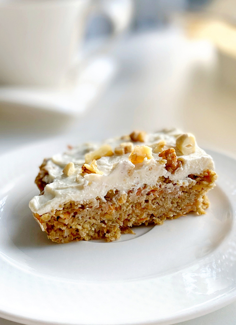 A slice of healthy low calorie carrot cake topped with chopped nuts, served on a white plate with a blurred background suggesting a cozy café ambiance.