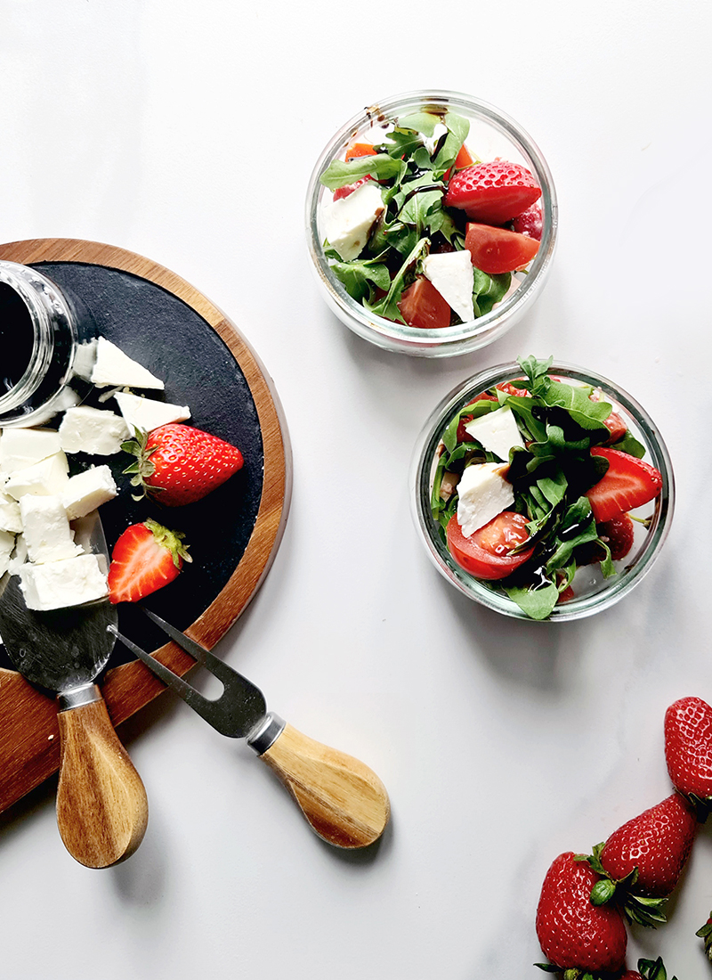 Top view of a fresh strawberry spring salad with arugula and feta cheese served on a wooden board with a jar of balsamic dressing and strawberries scattered around.