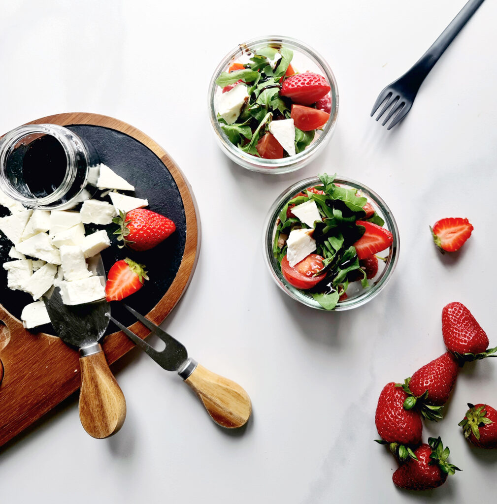 Top view of a fresh strawberry spring salad with arugula and feta cheese served on a wooden board with a jar of balsamic dressing and strawberries scattered around.