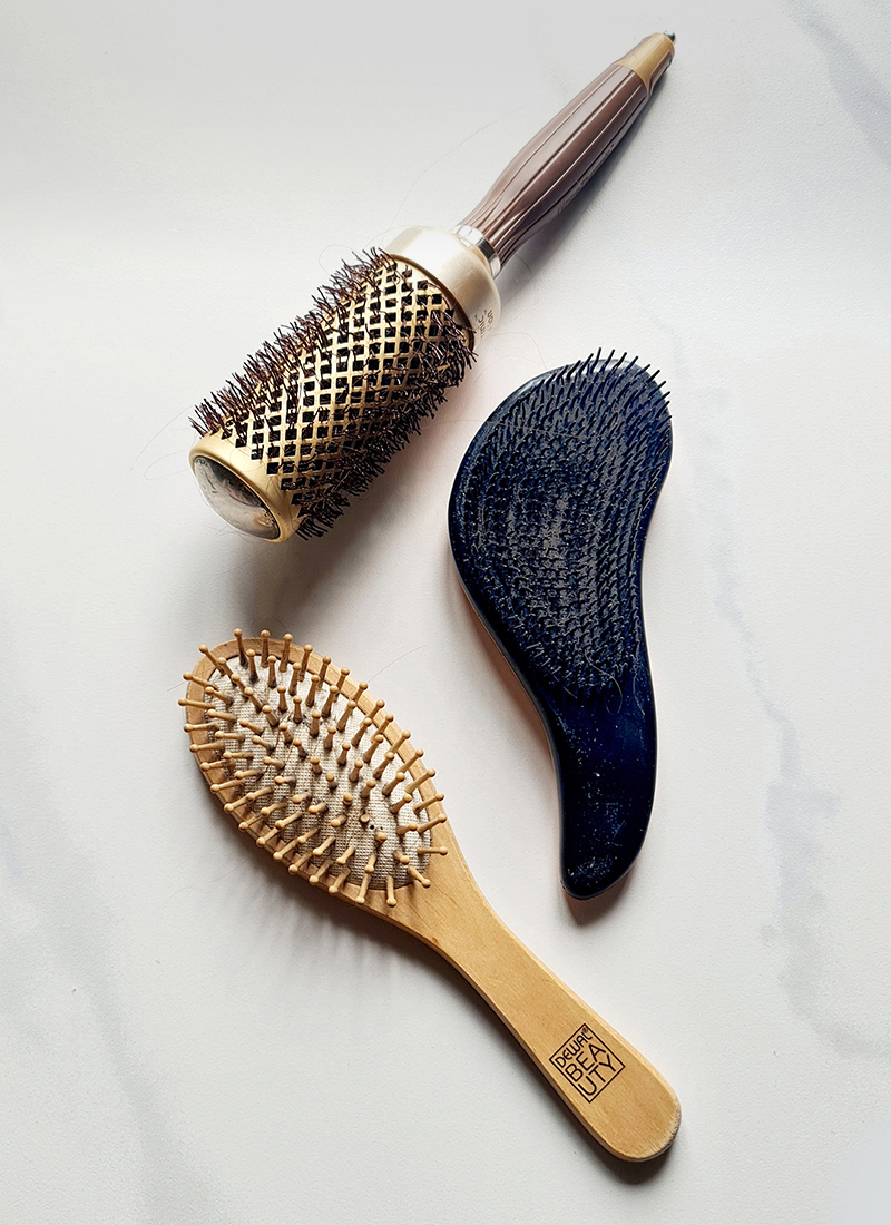 Three different types of dirty hairbrushes on a white background: a round brush with a metallic finish, paddle brush and a wooden brush with natural bristles.