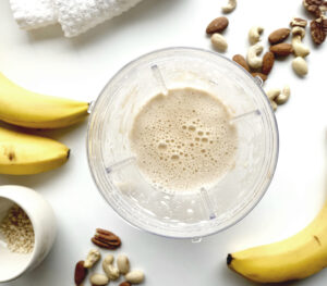 Top view of a blender containing a peanut butter banana smoothie, surrounded by bananas, nuts, and chopped almonds on a white surface.