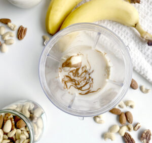 Top view of a blender with bananas, almond milk, and peanut butter for a peanut butter banana smoothie, surrounded by jars of nuts and whole bananas on a white surface.