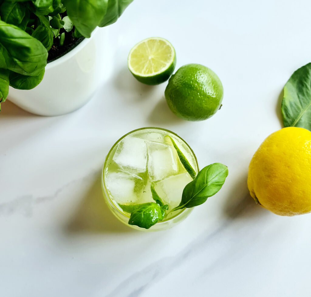 A top view of refreshing basil mocktail with ice and basil leaves in a glass, surrounded by fresh limes, a lemon, and more basil on a white surface.
