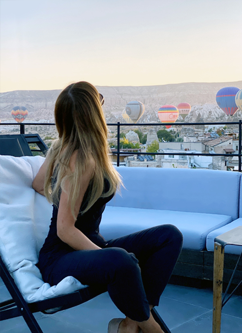 A woman sits on a rooftop terrace, looking at hot air balloons rising above a distant mountainous landscape, while reflecting on her journey of self-development and facing away from the camera.