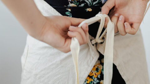 A woman with long hair wearing a floral dress ties the strings of a light-colored apron at the back, perhaps preparing to demonstrate some taste balancing techniques in cooking. The photo is taken from behind, focusing on the person's hands and the apron strings.