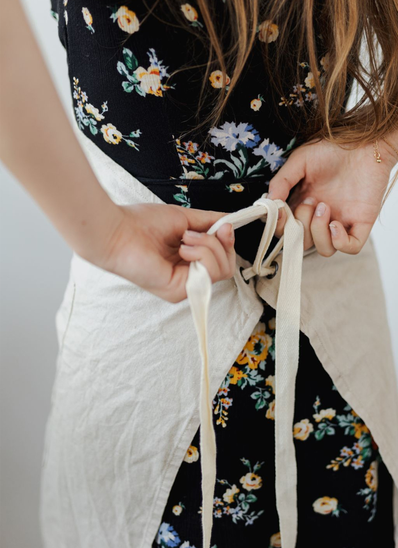 A woman with long hair wearing a floral dress ties the strings of a light-colored apron at the back, perhaps preparing to demonstrate some taste balancing techniques in cooking. The photo is taken from behind, focusing on the person's hands and the apron strings.