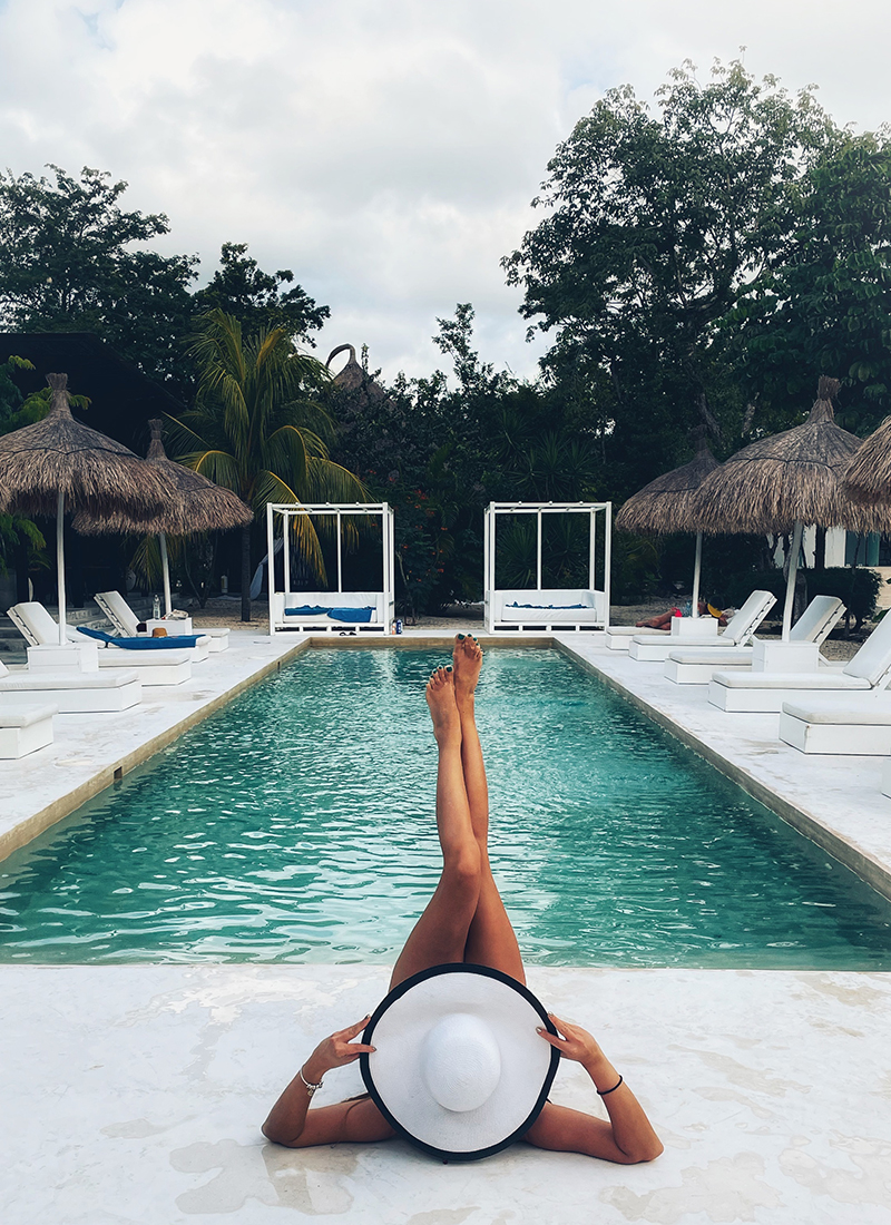 A woman lounging by a pool, legs raised with white hat, facing straw-roofed cabanas and lush greenery in Mexico. A serene and luxurious setting for relaxation.