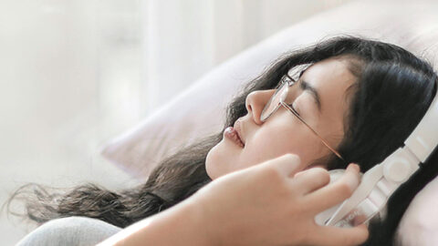 A young woman lies on a bed surrounded by soft pillows, listening to a guided meditation for sleep on white headphones, enjoying a serene moment bathed in natural light.