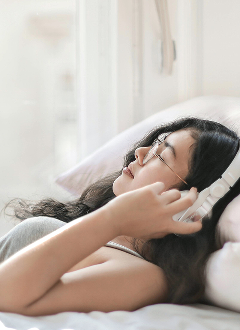 A young woman lies on a bed surrounded by soft pillows, listening to a guided meditation for sleep on white headphones, enjoying a serene moment bathed in natural light.