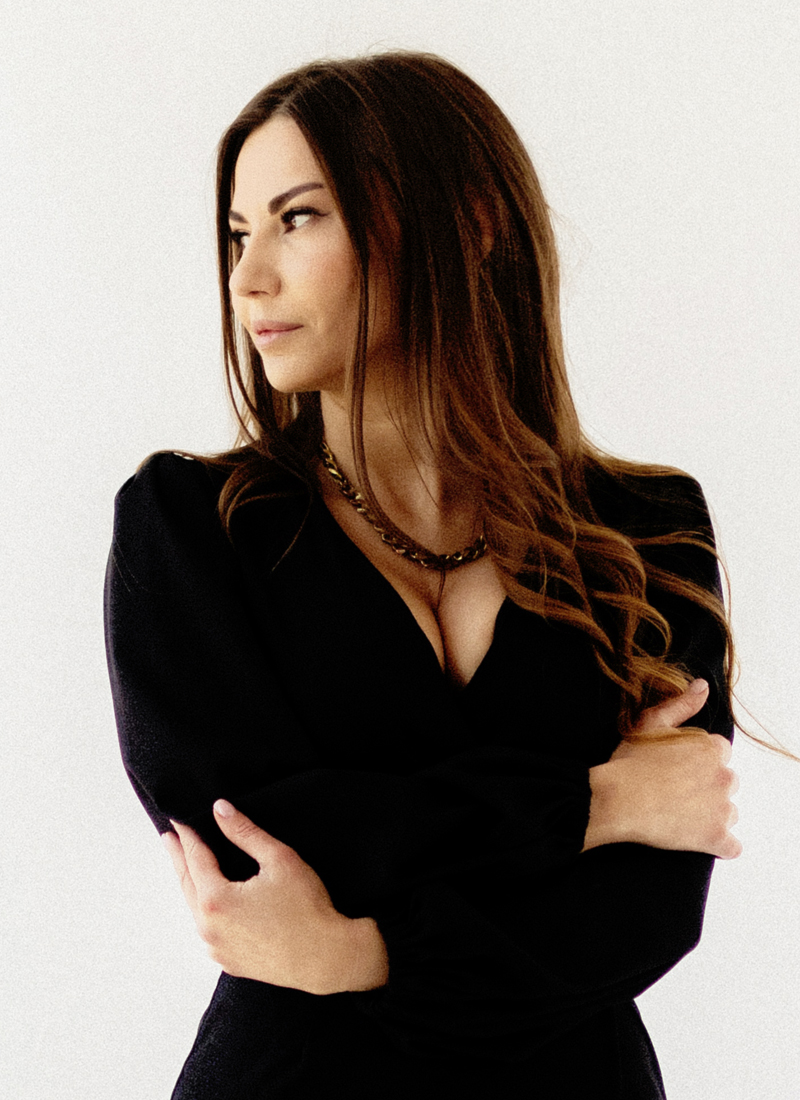 A woman in a chic black dress stands confidently against a white background, arms crossed, looking to her right, embodying women with high personal standards.