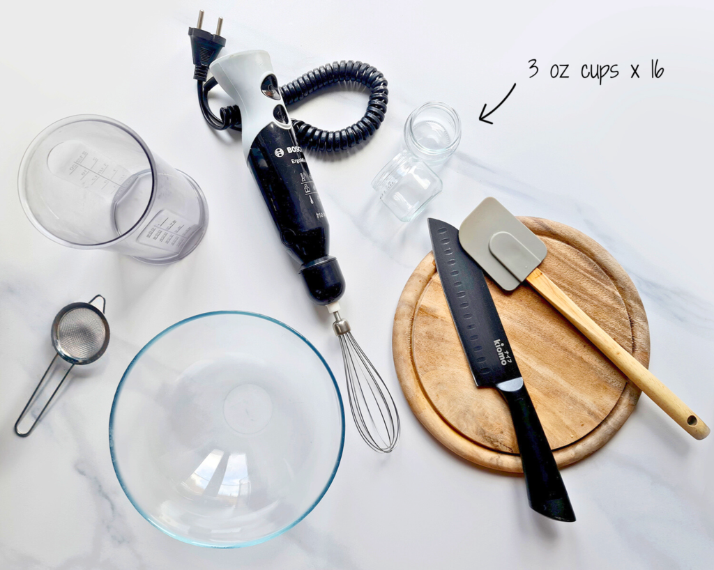 A kitchen setup includes a handheld mixer with a whisk attachment, a round glass bowl, a measuring cup, a black serrated knife, a grey spatula, and a wooden cutting board. Sixteen 3-ounce plastic cups—perfect for tiramisu dessert cups—are stacked in the background, with an arrow pointing to them.