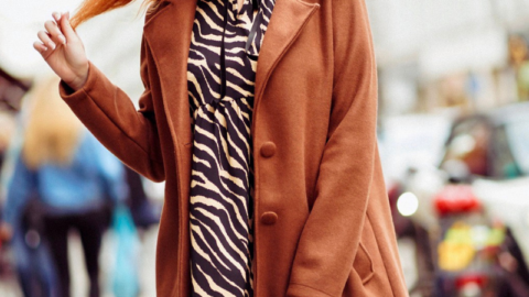 A stylish confident woman with red hair prepared for a photoshoot, walking confidently on a busy street wearing a tan coat over a zebra print dress, carrying a small blue handbag.