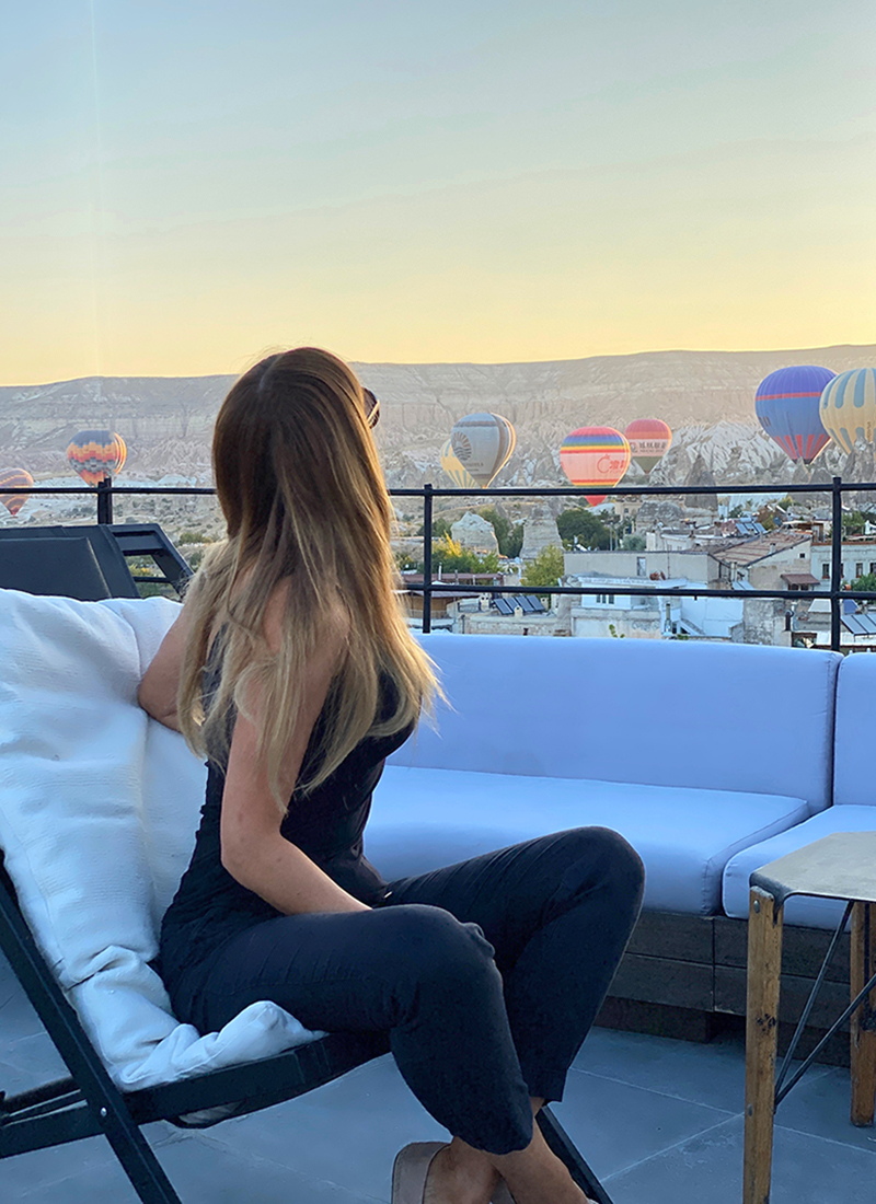A woman with long hair, wearing a black outfit, sits on a rooftop patio chair with her back to the camera. She contemplates an array of hot air balloons floating in the sky at sunrise or sunset, above a scenic landscape with hills and rooftops in the distance, embracing true goals amidst the tranquil scene.