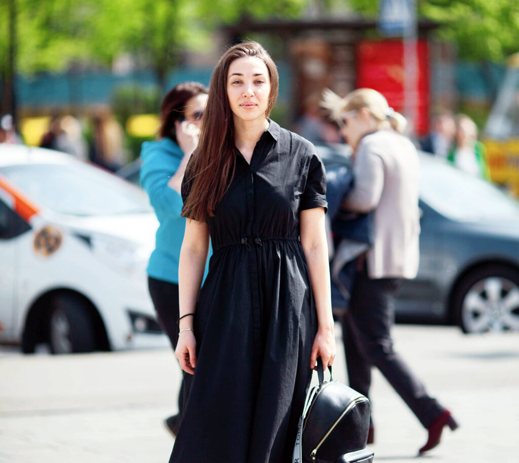 A woman with long brown hair wearing a black dress stands on a city street holding a black bag, perhaps pondering the best way to learn a language fast. Other pedestrians and parked cars are visible in the bright, sunny background.