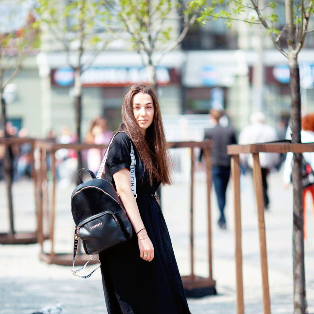 A young woman with long dark hair, wearing a black dress and carrying a black backpack, walks outdoors on a sunny day. There are young trees and blurred buildings in the background. The scene has a casual, urban feel with people and activity in the distance—perhaps discussing the fastest way to learn a language.
