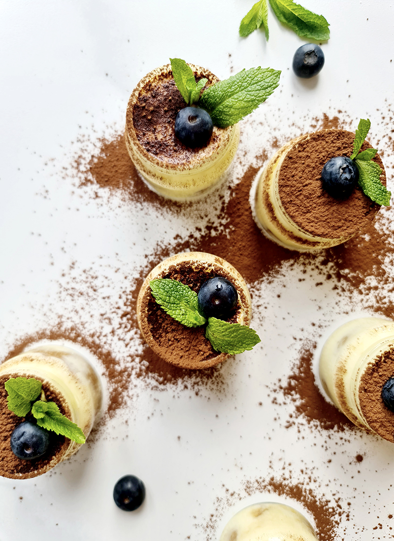 Overhead shot of beautifully presented tiramisu dessert cups garnished with blueberries and mint leaves. The desserts are dusted with cocoa powder and arranged on a white surface, creating a visually appealing contrast.