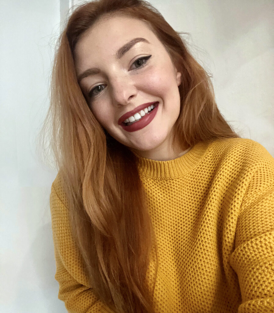 A smiling woman with long red hair, wearing a yellow knitted sweater, gently touches her head while posing for the camera. She has red lipstick and subtle makeup, highlighting a delicate smile.