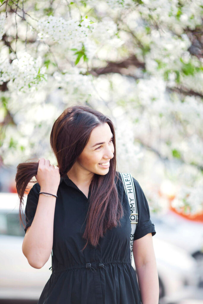 A woman with long, dark hair smiles while standing outdoors in front of a blooming tree with white flowers. She is wearing a black dress with a drawstring waist and has a white strap over her shoulder. As she contemplates how to learn a language fast, she holds a section of her hair and looks to the side.