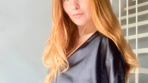 A woman with long red hair wearing a black elegant housecoat standing in a sunlit room, following her morning self-care routine, with white stairs and wall-mounted pictures in the background.