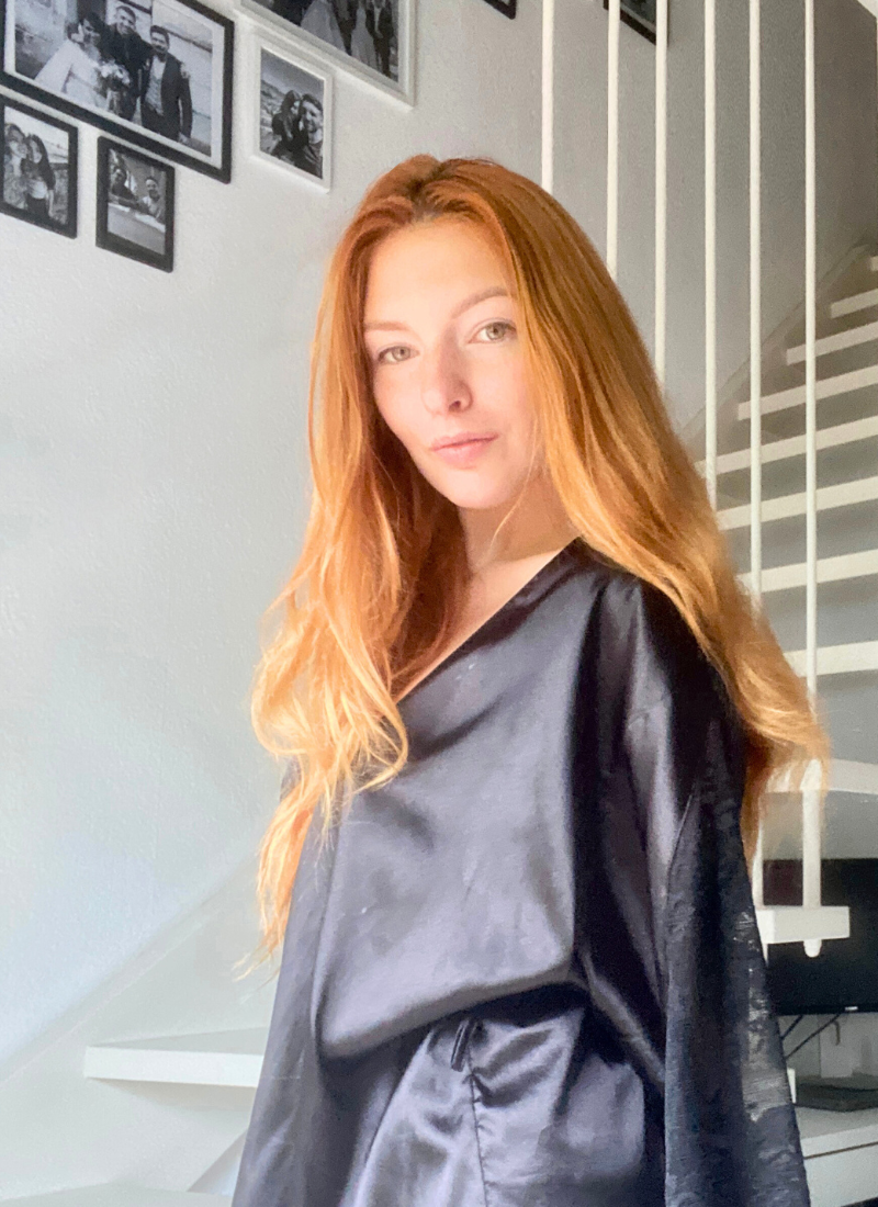 A woman with long red hair wearing a black elegant housecoat standing in a sunlit room, following her morning self-care routine, with white stairs and wall-mounted pictures in the background.