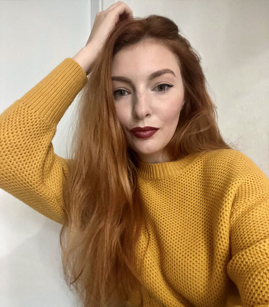 A woman with long red hair, wearing a yellow knitted sweater, gently touches her head while posing for the camera. She has red lipstick and subtle makeup, highlighting a delicate smile.