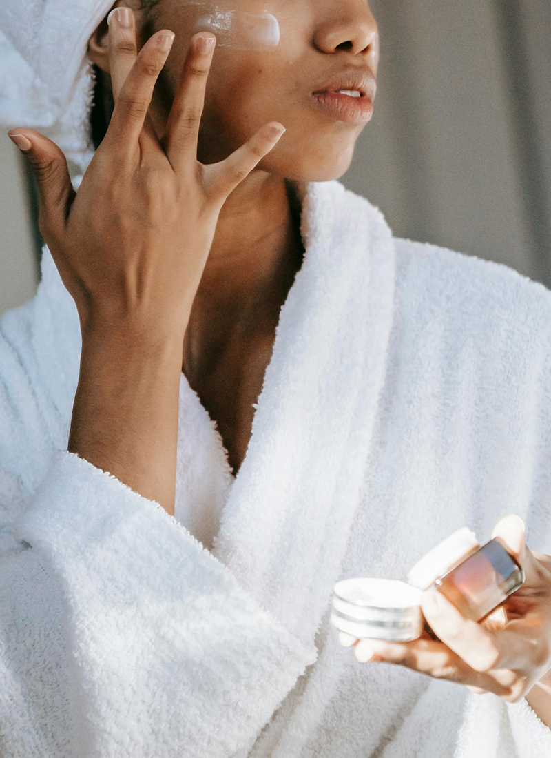 A person, immersed in their morning skin care routine, wears a white bathrobe and a towel wrapped around their head. They apply moisturizer to their face with one hand while holding a small jar of cream in the other hand. The background is softly lit, capturing the essence of tranquility.