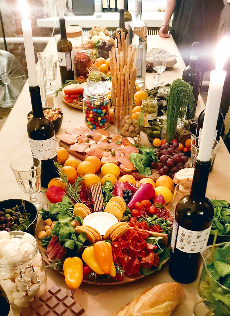 A table adorned with a variety of foods, including vegetables, fruits, chocolates, cheeses, breads, and snacks, surrounded by candles and bottles of wine. The colorful arrangement creates a festive and inviting atmosphere for a home party or gathering.