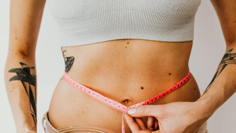 A woman bowl of salad with tomatoes in one hand while measuring her waist with a pink measuring tape with the other. With long, wavy blonde hair and several black tattoos on her arms, she exemplifies healthy nutrition habits against a plain background.