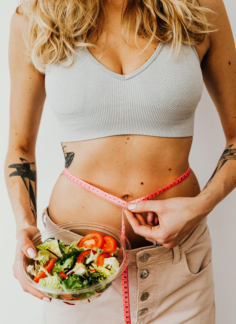 A woman bowl of salad with tomatoes in one hand while measuring her waist with a pink measuring tape with the other. With long, wavy blonde hair and several black tattoos on her arms, she exemplifies healthy nutrition habits against a plain background.