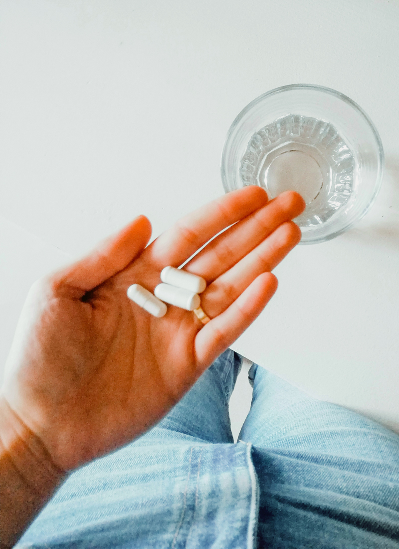 A hand holding three white capsules, recognized for their L-Lysine benefits, is extended over a clear glass of water. The person is wearing blue jeans.