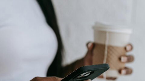 A person wearing a white shirt is holding a cup of coffee and a smartphone, presumably checking their task tracker app.