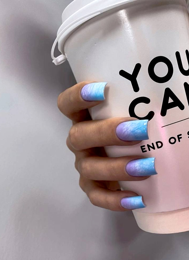 a hand with long nails holding a white coffee cup. The nails feature a blue-purple ombre summer nail idea design transitioning from light blue to purple with a glossy finish. The cup has text in black that reads, "YOU CAN.