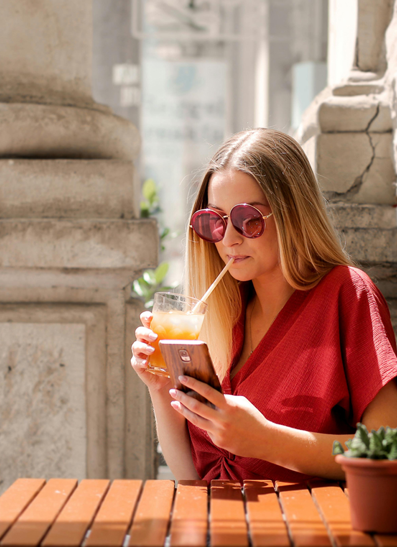 A woman in a red blouse and large round sunglasses is sitting at a wooden table outdoors. She is drinking a beverage with a straw and looking up the best apps for learning a new language on her smartphone. A small potted plant is on the table next to her. Background shows stone pillars and blurred scenery.