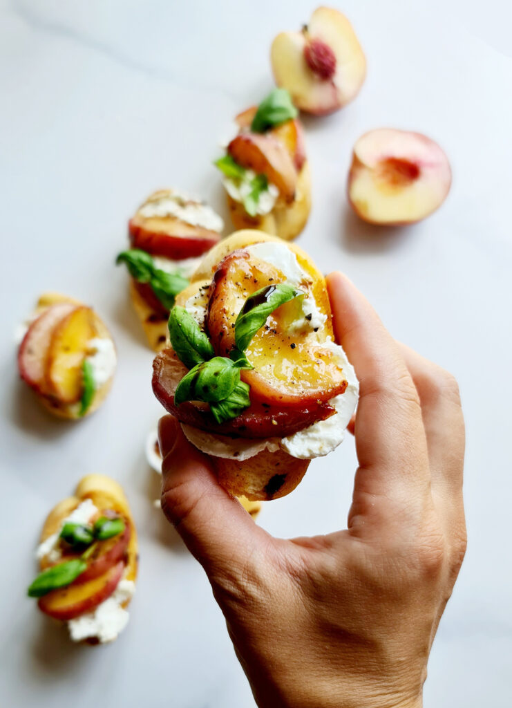 A hand holding a piece of peach bruschetta topped with burrata cheese, sliced peaches, fresh basil, and a drizzle of balsamic glaze. Other similar bruschettas and halved peaches are in the background on a white surface. This is the perfect appetizer idea for any gathering.