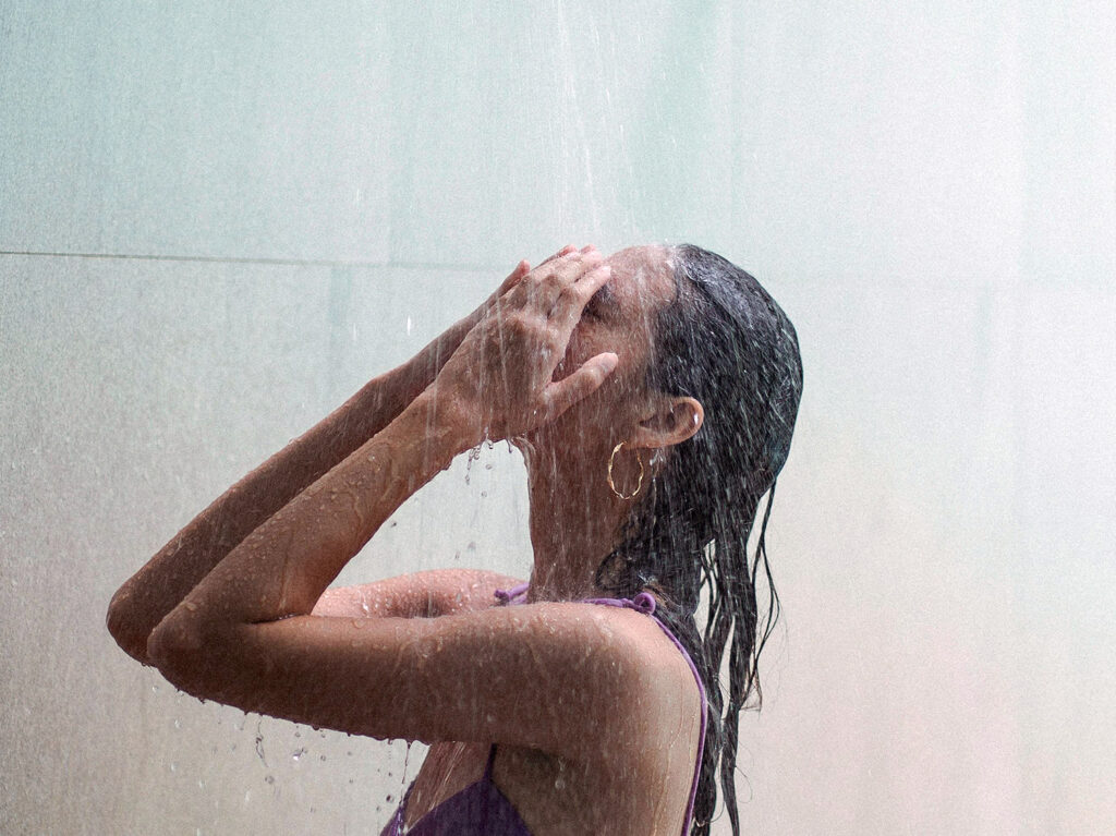 A person stands under a shower, wearing a purple tank top. Their face is obscured by their hands, placed on their forehead, as water streams down. They seem to be embracing the benefits of cold showers.