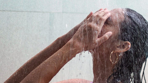 A person with wet hair stands under a shower, using both hands to cover their face. Water streams down onto their head and body, illustrating the benefits of cold showers. The person is wearing a purple sleeveless top and gold hoop earrings, set against a plain background.