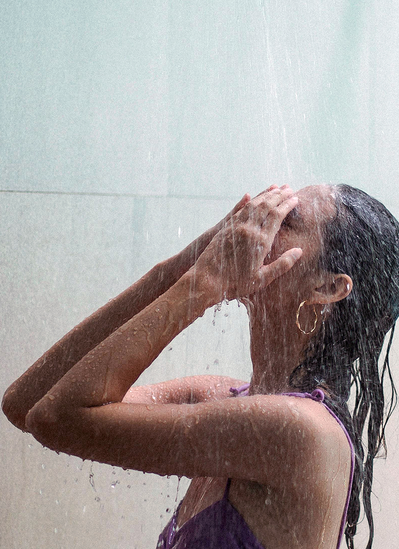 A person with wet hair stands under a shower, using both hands to cover their face. Water streams down onto their head and body, illustrating the benefits of cold showers. The person is wearing a purple sleeveless top and gold hoop earrings, set against a plain background.