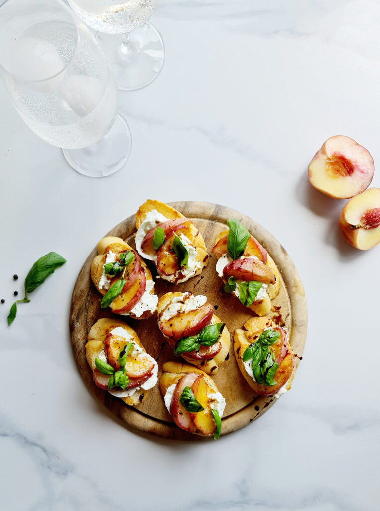 A wooden board holds several slices of toasted bread topped with creamy burrata, sliced peaches, fresh basil leaves, and a drizzle of balsamic glaze. This peach bruschetta's vibrant colors create an appetizing, gourmet presentation.